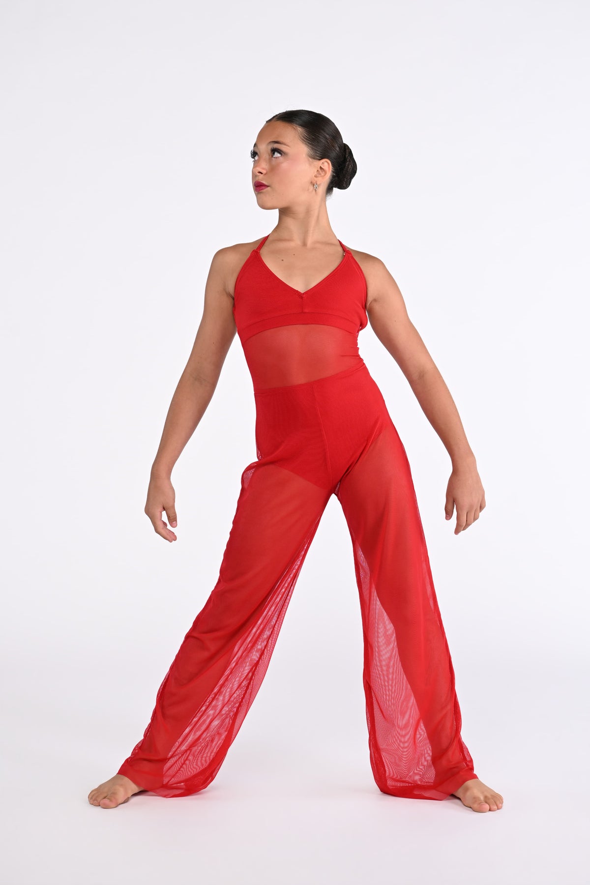 A woman modeling a bright red dance jumpsuit, featuring wide sheer mesh legs and crisscross halter-style straps, set against a plain white studio background.