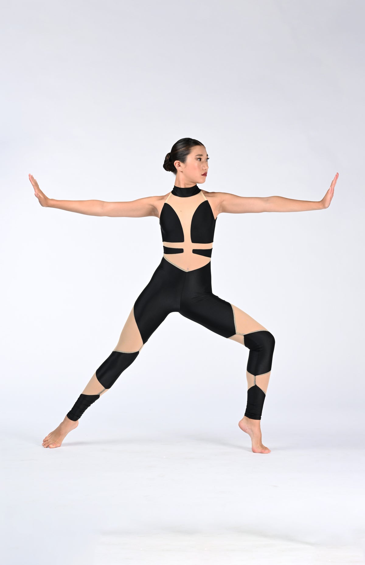 A woman in a black and beige cutout dance Unitard poses in a warrior II stance against a light gray studio background.